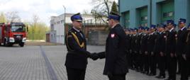 Dwóch strażaków podaje sobie rękę. Po prawej stronie strażacy ustawieni w dwuszeregu na tle budynku. W tle widać samochód strażacki oraz budynki.