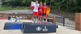 Trzech sportowców na podium podczas World Police & Fire Games w Birmingham. Zawodnik z Polski stoi po lewej stronie, trzyma polską flagę i medal. Obok niego na najwyższym stopniu podium stoi zawodnik w stroju Hiszpanii, również z medalem i flagą. Po prawej stronie trzeci medalista.