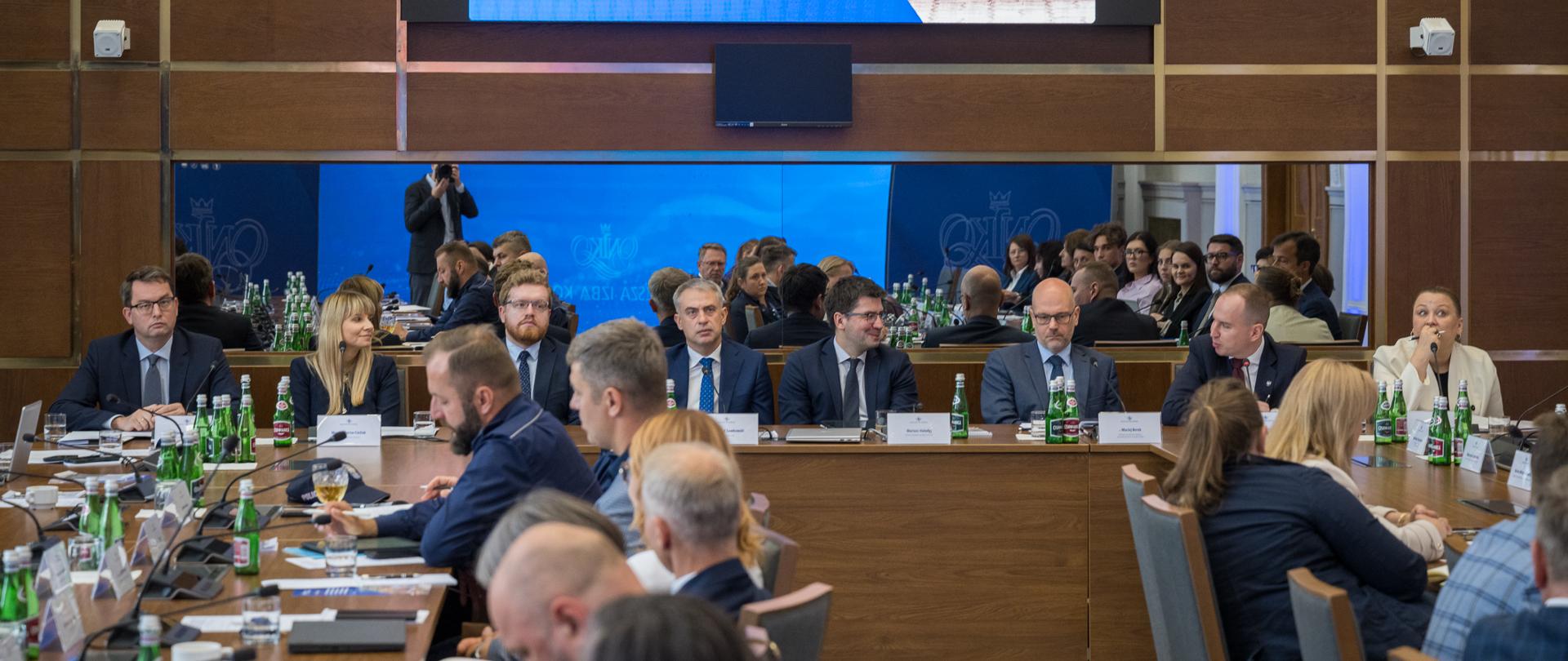 Zdjęcie przedstawia spotkanie w sali konferencyjnej Najwyższej Izby Kontroli (NIK). Grupa ekspertów i uczestników w strojach biznesowych siedzi przy długim, drewnianym stole wyposażonym w mikrofony, laptopy i napoje. Na ścianie wisi duży ekran z prezentacją pt. „Panel ekspertów: Bezpieczne dziecko w świecie cyfrowym – systemowe rozwiązania, wspólna odpowiedzialność”, z datą 8 lipca 2026 r. Na slajdzie widoczne jest zdjęcie dzieci korzystających ze smartfonów.