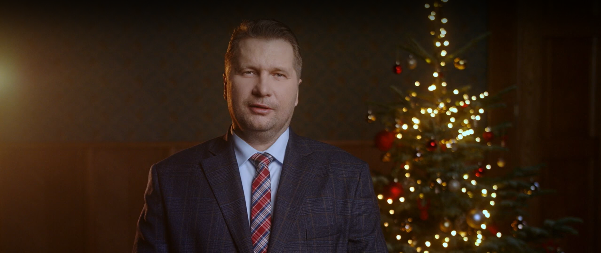 Minister Przemysław Czarnek ubrany w garnitur i krawat w kratkę patrzy w obiektyw. W tle choinka z kolorowymi bombkami i światełkami 