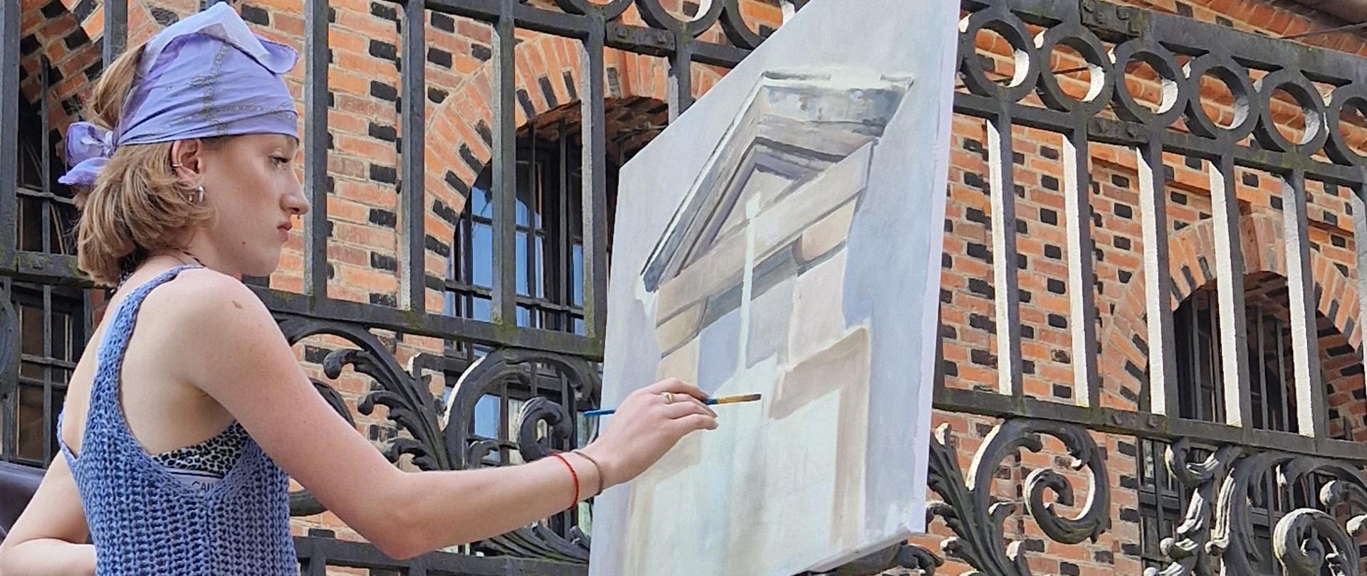 Na zdjęciu znajdują się dwie postacie. Od lewej strony pani Magdalena Wanat-Drobisz ubrana w turkusową sukienkę i jasny kapelusz. Nauczyciel ma prawą rękę wyciągniętą ku górze, w ten sposób pokazuje na co trzeba zwrócić uwagę malując obraz. Po prawej stronie znajduje się uczennica malująca obraz. Obraz ustawiony jest na plenerowej sztaludze. Postacie stoją na betonowym chodniku. Tuż za postaciami znajduje się kute metalowe ogrodzenie usytuowane na murze wykonanym z czerwonej cegły. W tle widzimy stare miasto w Sandomierzu 