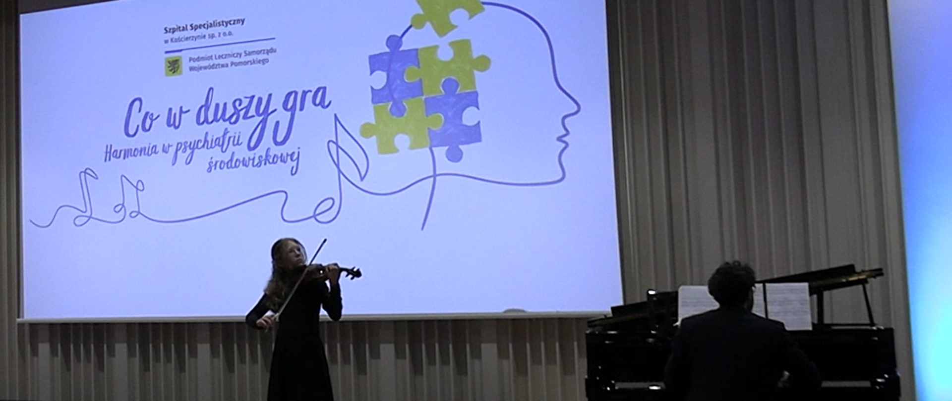 Na zdjęciu na scenie dziewczyna- brunetka ubrana na czarno, która gra na skrzypcach. Po prawej stronie zdjęcia przy czarnym fortepianie mężczyzna- brunet w czarnej marynarce. Na ścianie wyświetla się niebieski napis "co nam w duszy gra. Harmonia w psychiatrii środowiskowej". Po prawej stronie wyświetlanego obrazu puzzle żółte i niebieskie na grafice ludzkiej głowy. 