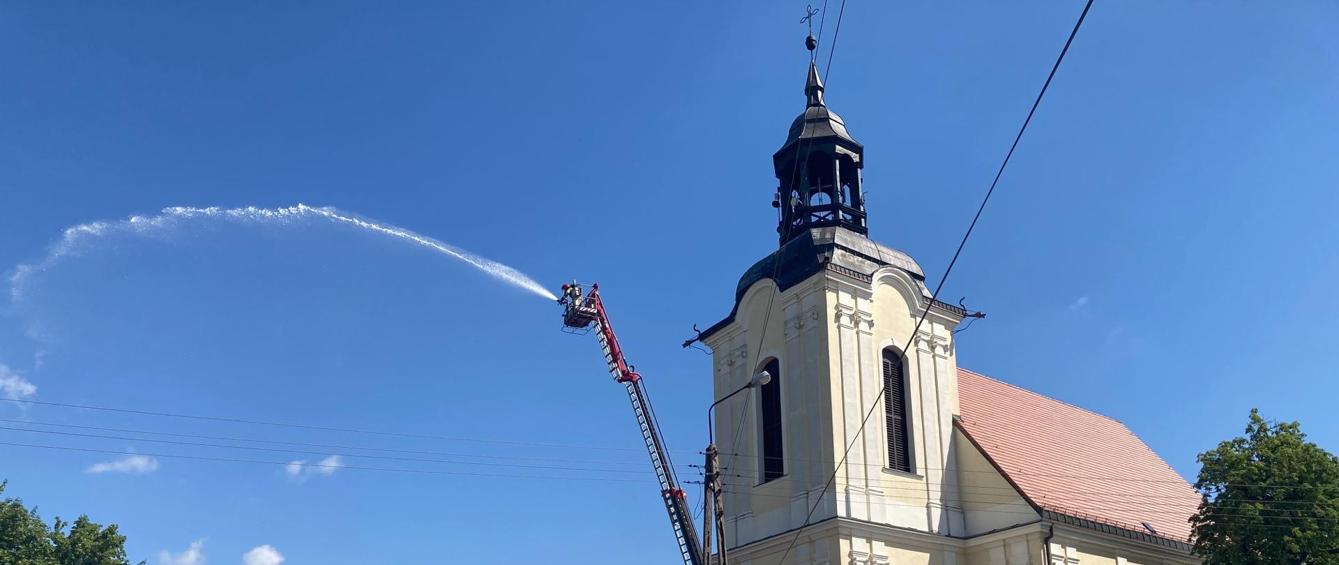 Zdjęcie wykonano w ciągu dnia przed budynkiem kościoła w miejscowości w Zamartem. Przedstawia ćwiczenia obiektowe z użyciem podnośnika hydraulicznego, kilkoro strażaków Ochotniczej Straży Pożarnej stoi przy samochodzie ratowniczo- gaśniczym. Na dalszym planie bliżej kościoła stoi rozstawiony podnośnik hydrauliczny z koszem z którego strażak Państwowej Straży Pożarnej podaje prąd wody na pobliskie drzewa. 