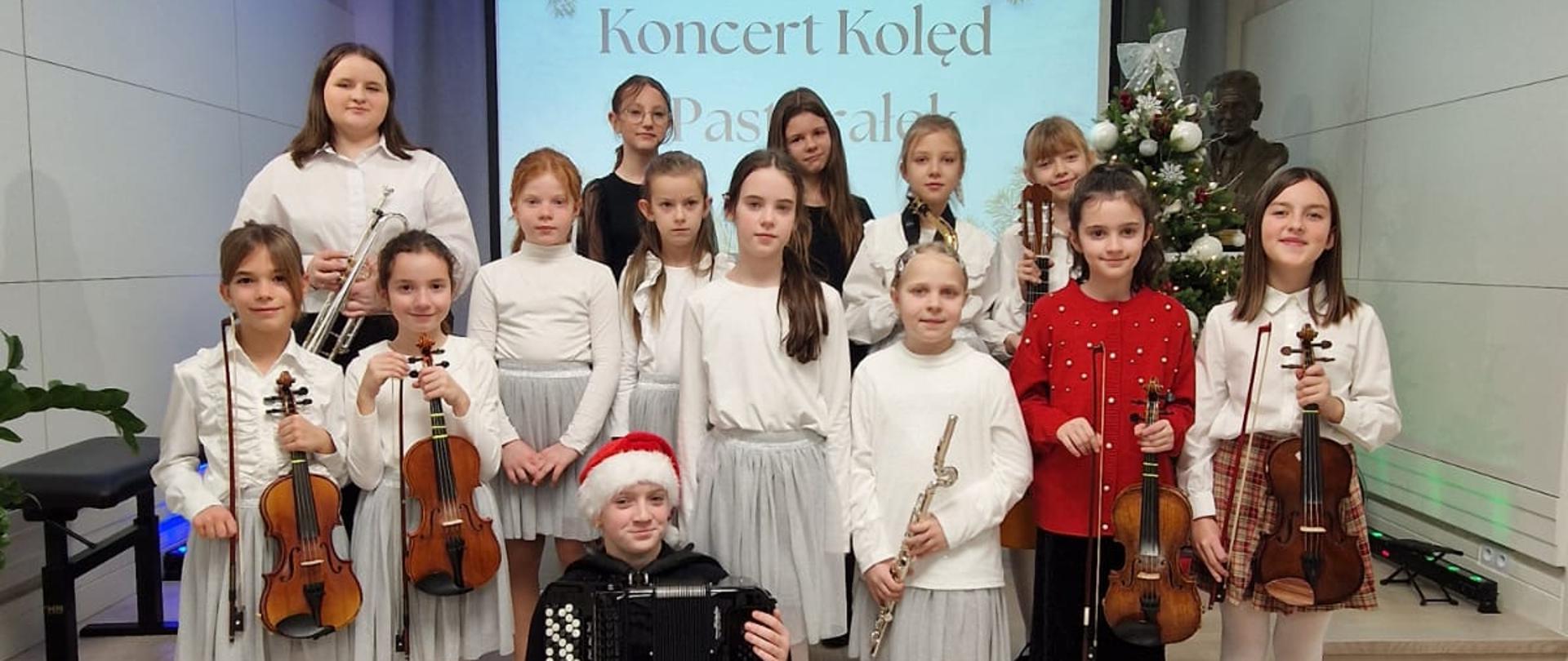 Zdjęcie przedstawia grupę uczniów ubranych w odświętne, białe i jasnoszare stroje, niektóre trzymają instrumenty takie jak skrzypce, flet poprzeczny, trąbkę czy gitarę. W pierwszym rzędzie, na dolnym stopniu sceny, siedzi dziewczynka z akordeonem ubrana w czarny strój i czerwoną czapkę Mikołaja. W tle widoczny jest duży ekran z napisem "Koncert Kolęd i Pastorałek" ozdobiony grafiką świąteczną z jemiołą i bombkami. Obok ekranu stoi udekorowana choinka oraz popiersie patrona szkoły. 