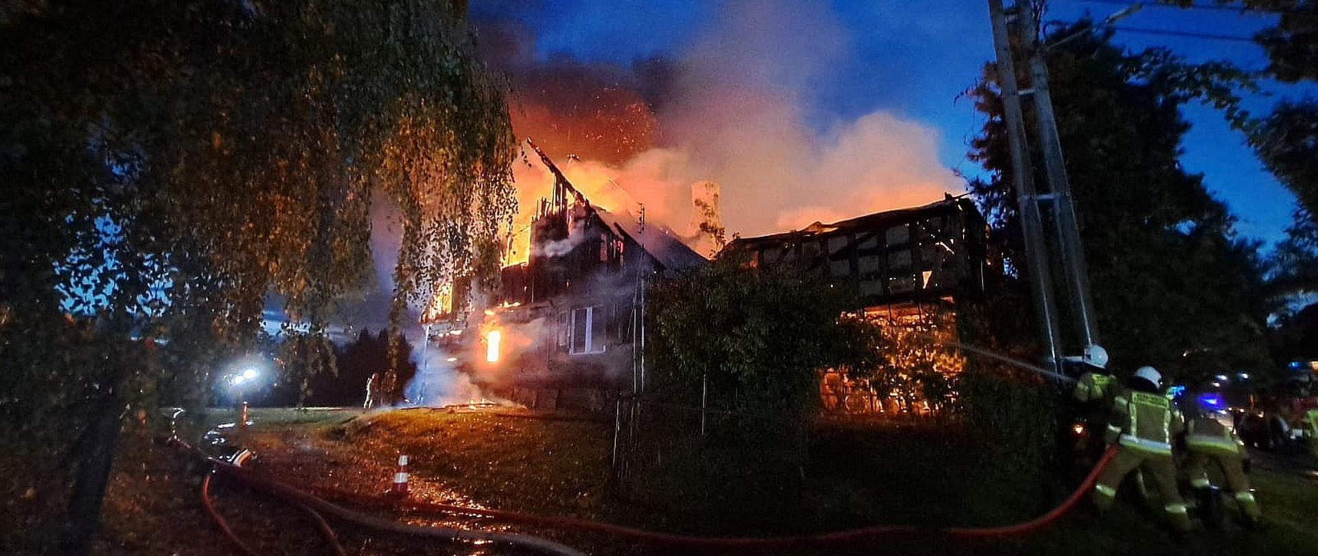 W tle palący się nocą budynek mieszkalnych. Dom w całości jest objęty ogniem. Z prawej strony widoczne sylwetki strażaków. Strażacy podają prąd wody na palący się obiekt.