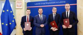 Wojewoda lubelski Krzysztof Komorski i trzech mężczyzn w garniturach stoją na tle flagi unijnej i granatowej ścianki z napisem Lubelski Urząd Wojewódzki w Lublinie.