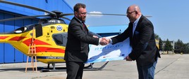 Minister zdrowia Łukasz Szumowski przekazał symboliczny czek dyrektorowi Lotniczego Pogotowia Ratunkowego Robertowi Gałązkowskiemu. W tle żółty helikopter Lotniczego Pogotowia Ratunkowego.
