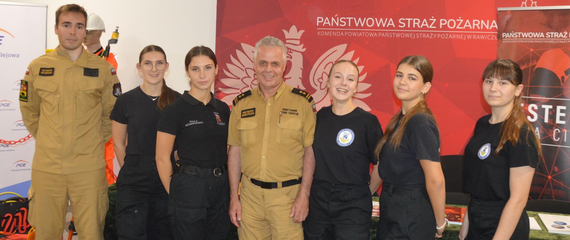 Noc zawodowców. Zdjęcie przedstawia korytarz w Zespole Szkół Zawodowych w Rawiczu. Na tle ścianki reklamowej KP PSP w Rawiczu do wspólnego zdjęcia w towarzystwie dwóch strażaków PSP pozują uczennice klasy mundurowej jednej ze szkół na terenie powiatu. Dziewczyny mają na sobie mundury w kolorze czarnym.