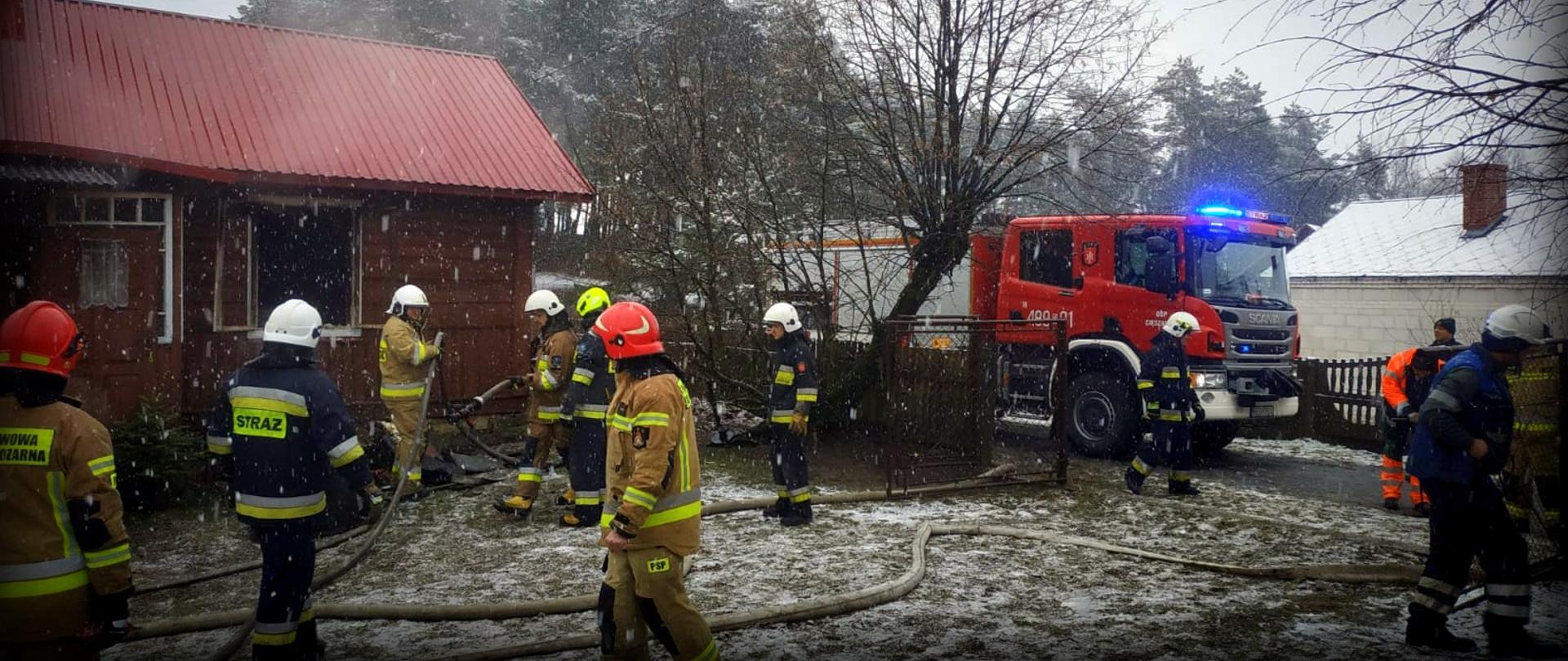Zdjęcie zrobione na zewnątrz. Strażacy w trakcie działań ratowniczo-gaśniczych. W tle widać drewniany dom. Z okien domu unosi się dym. Po prawej stronie stoi strażacki samochód ratowniczo-gaśniczy.