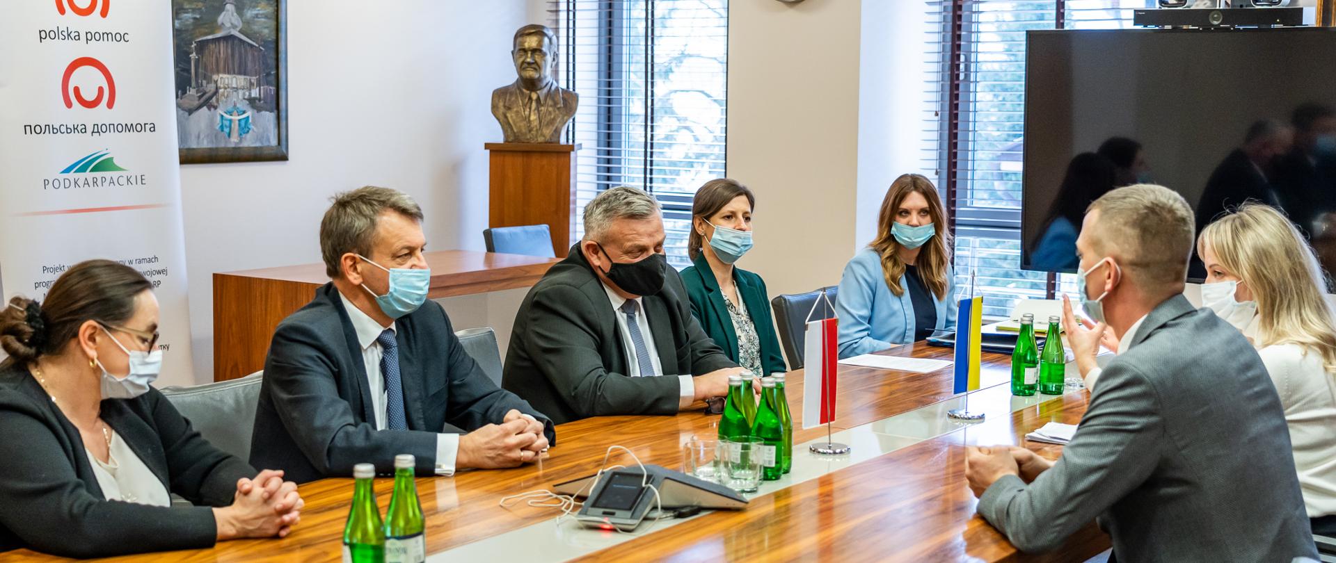 Ludzie siedzący po obydwu stronach stołu. Na stole ustawione są dwie małe flagi Polski i Ukrainy oraz butelki z wodą.