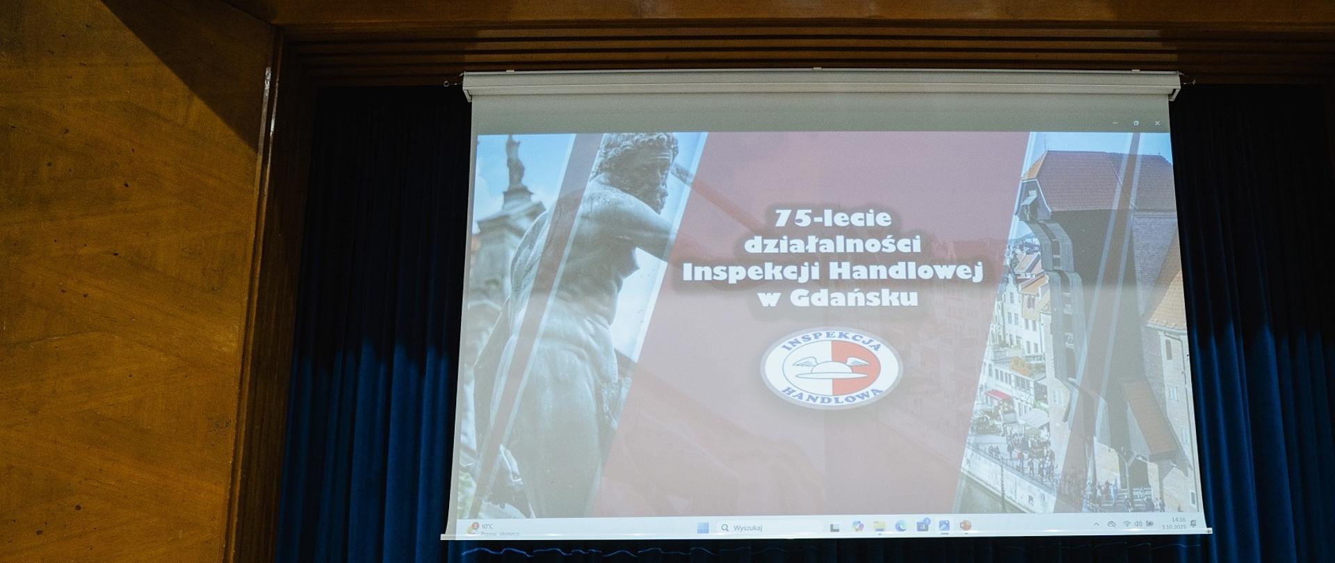 Telebim z wyświetlonym napisem 75-lecie działalności Inspekcji Handlowej w Gdańsku w tle granatowa kurtyna.