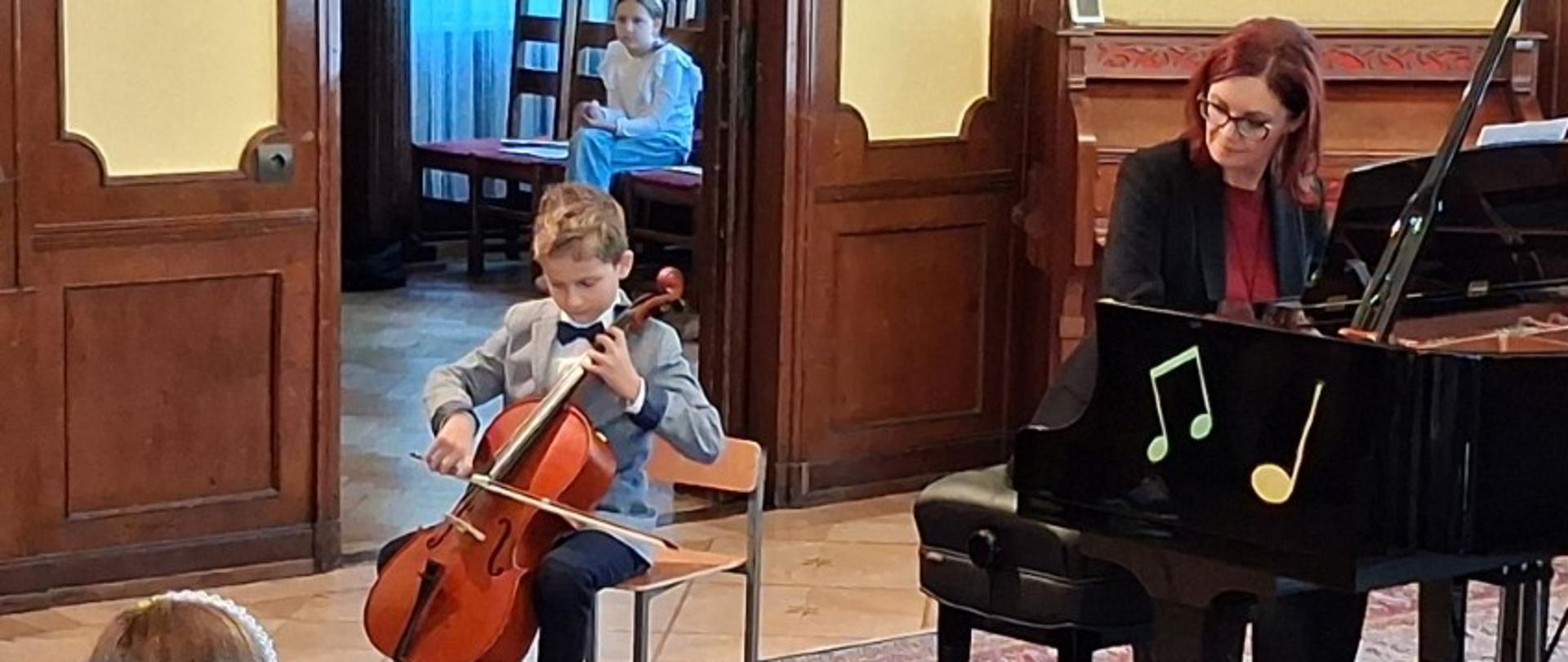 Zdjęcie kolorowe chłopiec w jasnej, szarej marynarce i spodniach siedzi na krześle, grając na wiolonczeli. Po prawej stronie siedzi kobieta w czarnej marynarce i czerwonej bluzce, grając na czarnym fortepianie, udekorowanym kolorowymi nutkami. W głębi otwarte drzwi, za którymi widać dziewczynkę siedzącą na krześle. Na pierwszym planie widoczne głowy publiczności.
