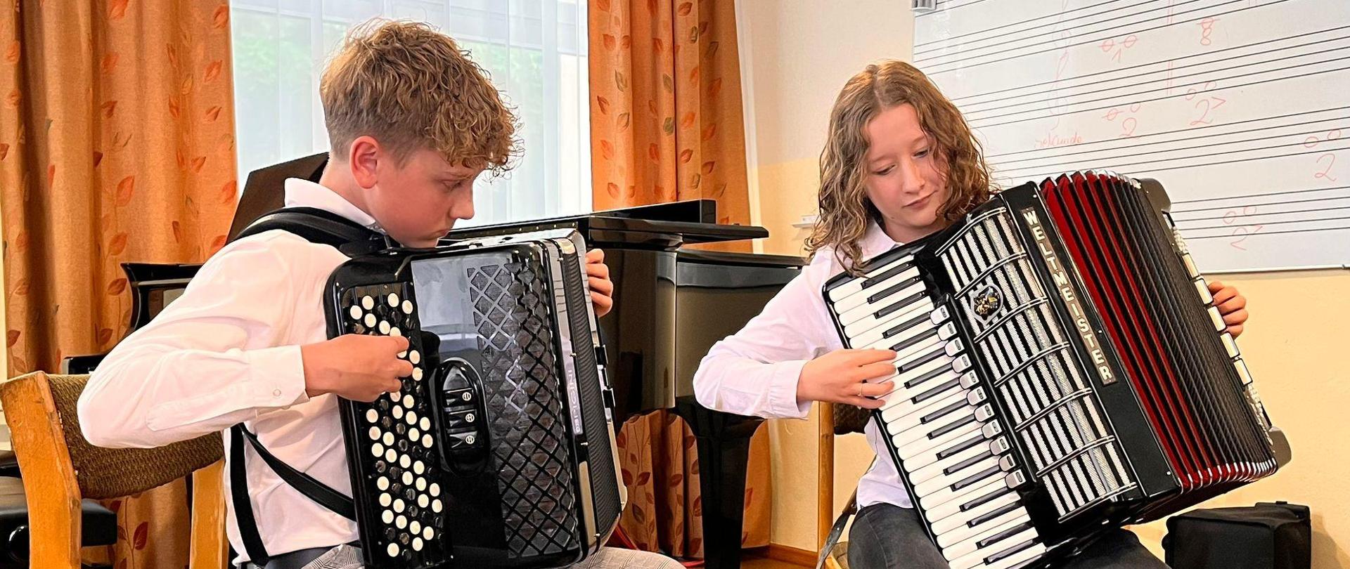 Sala lekcyjna. Duet akordeonowy. Na krześle siedzą i grają na akordeonie chłopiec po lewej stronie i dziewczynka po prawej. Na ścianie tablica, na środku czarny fortepian na ziemi podnóżek. Głęboko w tle okno z dużymi pomarańczowymi zasłonami.