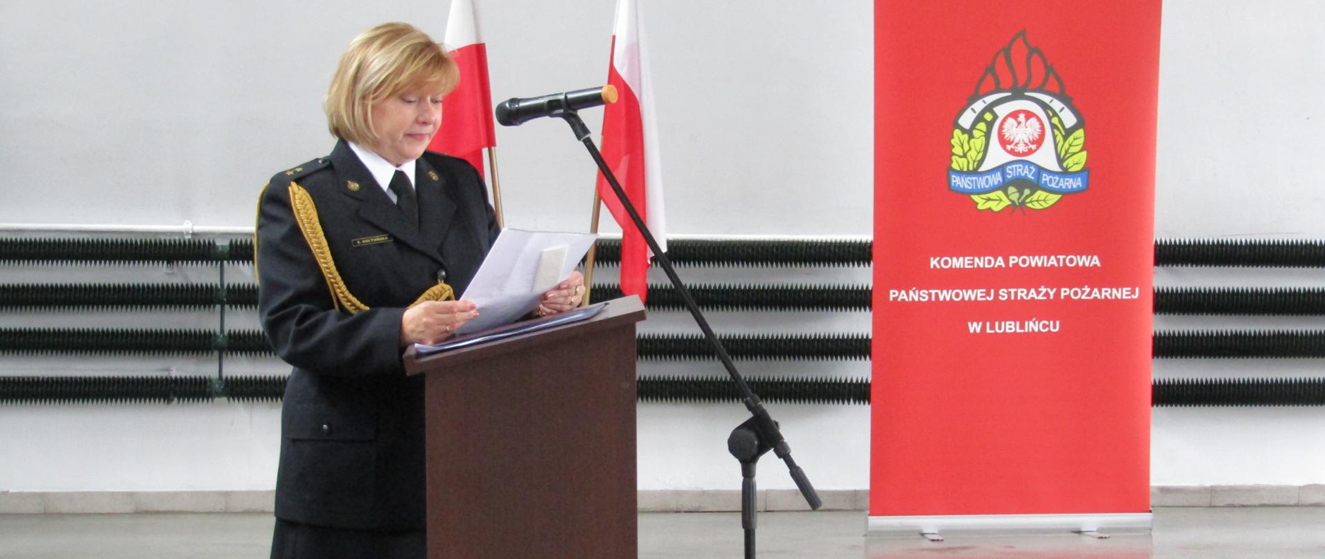 Zdjęcie zrobione w pomieszczeniu strażnicy na tle baneru Komendy Powiatowej PSP w Lublińcu oraz Flag Państwowych. Na pierwszym planie funkcjonariusz odchodzący na zaopatrzenie emerytalne, w umundurowaniu galowym, przemawia do mikrofonu stojącego przy mównicy.
