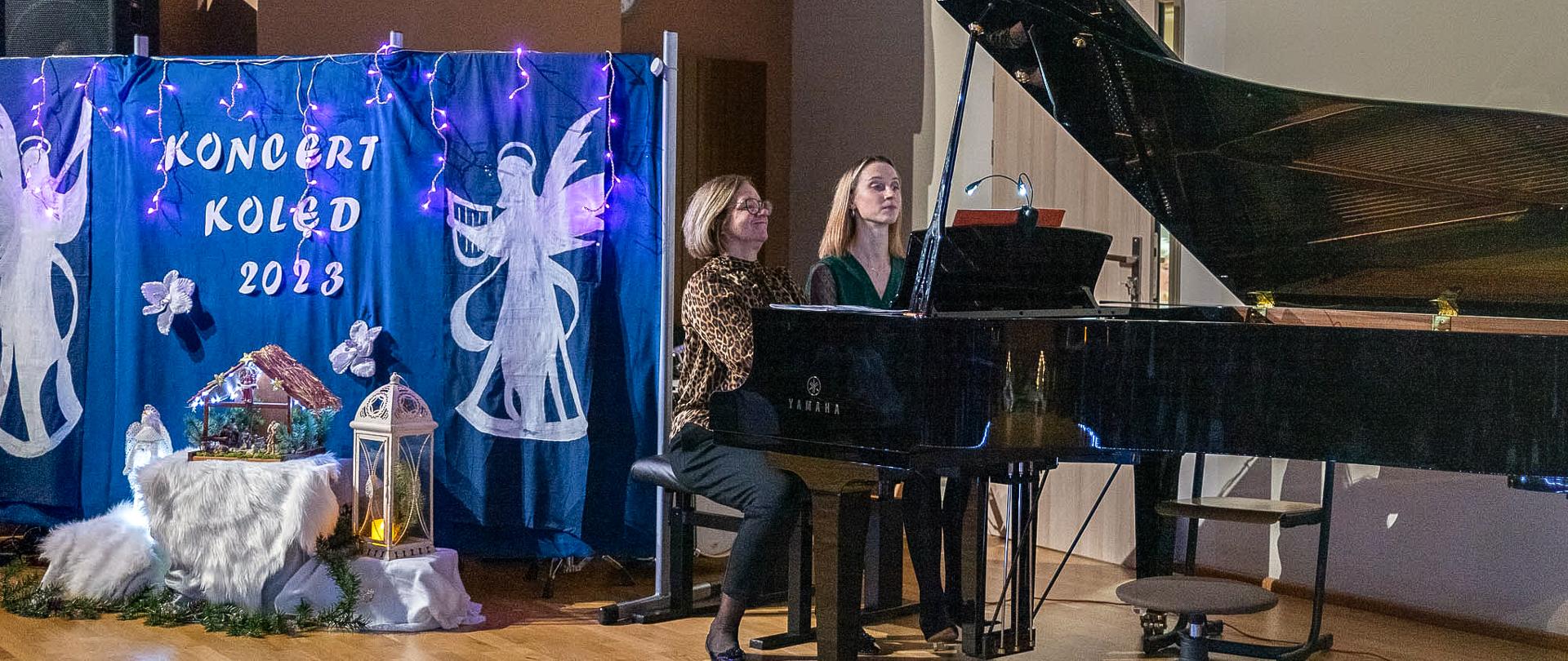 Na zdjęciu w centralnej części kadru dwie grające pianistki na cztery ręce. Są to nauczycielki gry na fortepianie - Beata Felczykowska oraz Julianna Bartyzel. Po lewej stronie kadru na niebieskim tle duży napis "Koncert kolęd 2023" a pod spodem żłóbek. 