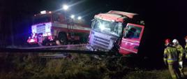 Zdjęcie przedstawia samochód ciężarowy, który wjechał do rowu. Zdjęcie zrobione porą nocną. Widać uszkodzone barierki energochłonne oraz uszkodzoną kabinę pojazdu. Na drugim planie widać samochód straży pożarnej oświetlający teren wypadku. Po prawej stronie widać pracujących strażaków. 