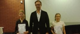 Na zdjęciu widzimy trzy osoby stojące na tle jasnego ekranu projekcyjnego i ściany z czerwonej cegły. Pośrodku znajduje się dorosły mężczyzna w okularach, ubrany elegancko w ciemną marynarkę, białą koszulę i spodnie w kolorze beżowym. Po jego lewej (z naszej perspektywy prawej) stronie stoi młoda dziewczynka z długimi, ciemnymi włosami spiętymi w kucyk, ubrana w jasną koszulkę. Trzyma w rękach dyplom oraz torbę. Po prawej stronie mężczyzny (z naszej perspektywy lewej) stoi druga dziewczynka, o jasnych, kręconych włosach, ubrana w ciemną bluzkę z wzorem. Ona również trzyma w rękach dyplom i torbę. W tle widoczny jest fragment fortepianu i grzejnik. Zdjęcie zostało wykonane prawdopodobnie podczas uroczystości wręczenia nagród lub dyplomów, najpewniej w szkole muzycznej lub podobnej instytucji edukacyjnej.