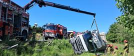 Zdjęcie przedstawia wypadek ciągnika siodłowego z naczepą. Jest pora dzienna, słoneczna pogoda. Na zdjęciu widać wywrócony pojazd który leży na bok w pobliskim rowie. Obok pojazdu klęczą strażacy ubranych w ubrania specjalne którzy wykonują dostęp do osoby poszkodowanej. Kabina pojazdu jest zapięta do żurawia ciężkiego samochodu ratownictwa drogowego. Po lewej stronie widać czerwone pojazdy pożarnicze i karetkę Zespołu Ratownictwa Medycznego.