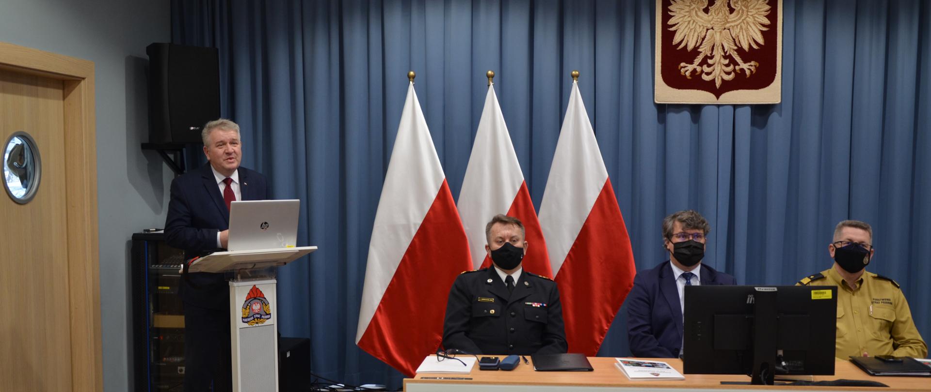 Zdjęcie z narady, na lewo przy mównicy stoi Wicewojewoda Mazowiecki Sylwester Dąbrowski, przy stole z prawej strony zdjęcia siedzą Mazowiecki Komendant Wojewódzki PSP st. bryg. Jarosław Nowosielski, Sekretarz Stanu w Ministerstwie Spraw Wewnętrznych i Administracji Maciej Wąsik, Zastępca Komendanta Głównego PSP nadbryg. Krzysztof Hejduk oraz Wicewojewoda Mazowiecki Sylwester Dąbrowski.