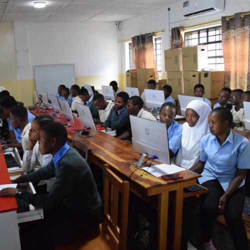 Młodzież siedząca w ławkach przed ekranami komputerów.