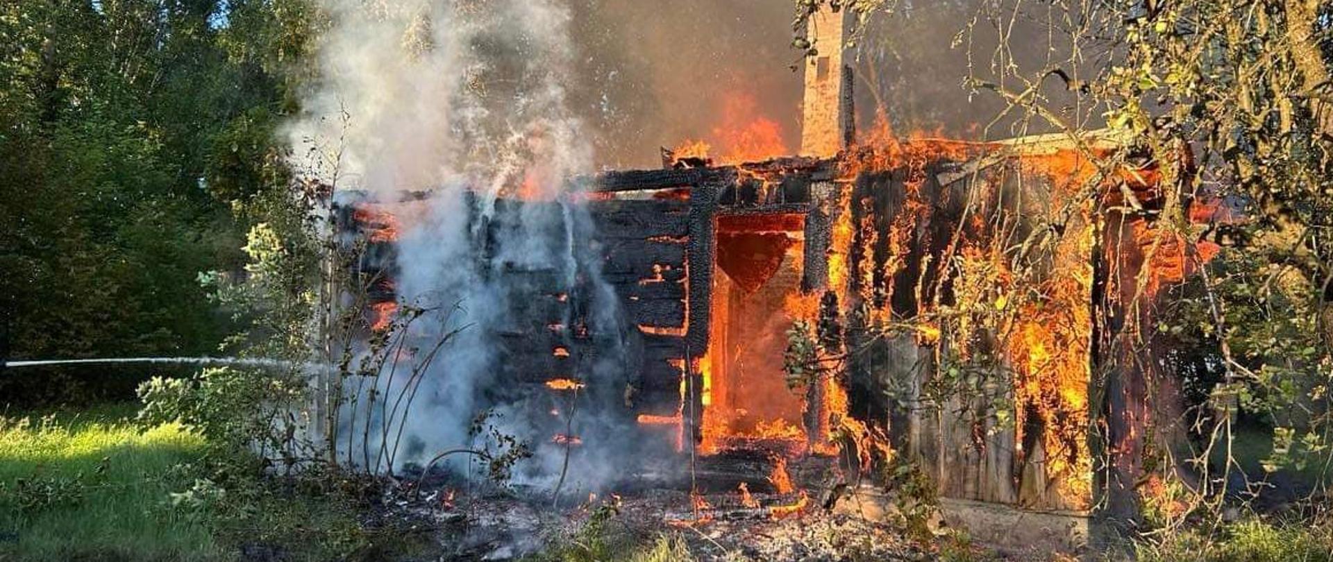 Na zdjęciu widać budynek objęty silnym pożarem w ciągu dnia. Płomienie intensywnie trawią jego konstrukcję, a gęsty dym unosi się w górę. Budynek wydaje się być wykonany z drewna lub podobnych materiałów łatwopalnych, a znaczna jego część jest zwęglona na czarno, szczególnie po lewej stronie, gdzie widoczny jest strumień wody z węża, co wskazuje na trwające działania gaśnicze. Jaskrawo pomarańczowe i żółte płomienie wyraźnie widać po prawej stronie budynku, trawiąc drewniane ściany. Dym, w kolorze od białego do ciemnoszarego, unosi się w górę z różnych części płonącego budynku. W tle widoczne są zielone drzewa i liście, a po prawej stronie – czyste, błękitne niebo. Na pierwszym planie widać zieloną trawę i zwęgloną ziemię, co wskazuje na rozprzestrzenianie się ognia. Całość obrazu oddaje skalę zniszczeń spowodowanych przez pożar.