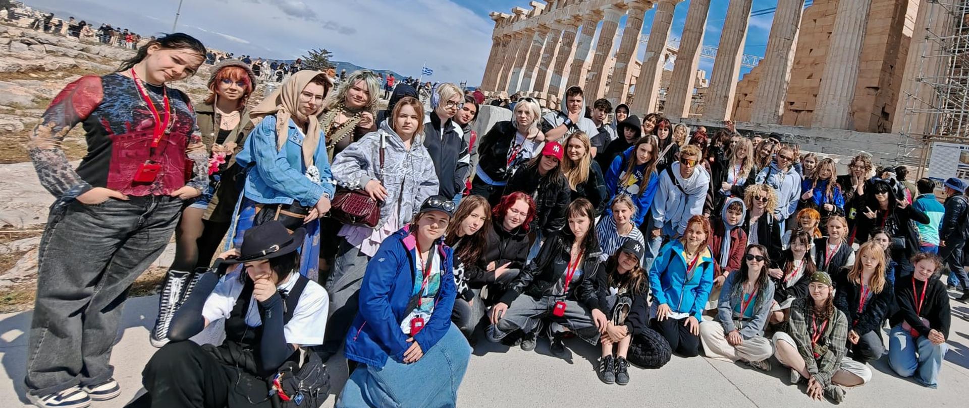 Na zdjęciu widzimy grupę uczniów i nauczycieli biorących udział artystycznej wyprawie do Grecji. Fotografia autorstwa Katarzyny Gwizdała-Kłak została wykonana na wzgórzu Akropolu. W tle widzimy świątynię Partenon.