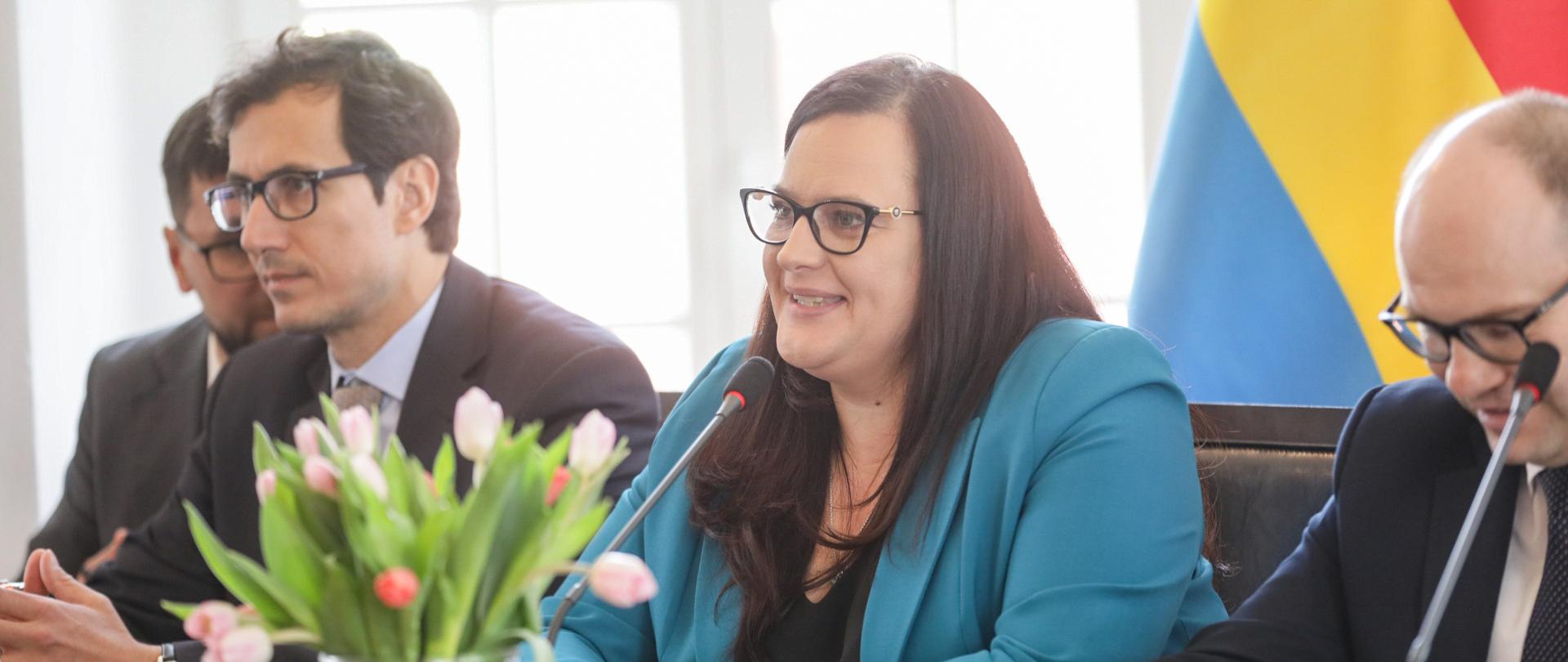 Trzy osoby siedzą przy stole. W środku jest wiceminister Małgorzata Jarosińska-Jedynak. Z przodu na stole stoi wazon z tulipanami. 