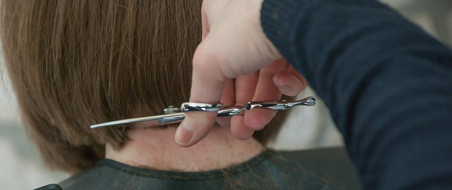 włosy kobiety do ramion strzyżone w salonie fryzjerskim. Widoczna ręka z nożyczkami i kawałek grzebienia, klientka siedzi tyłem. 