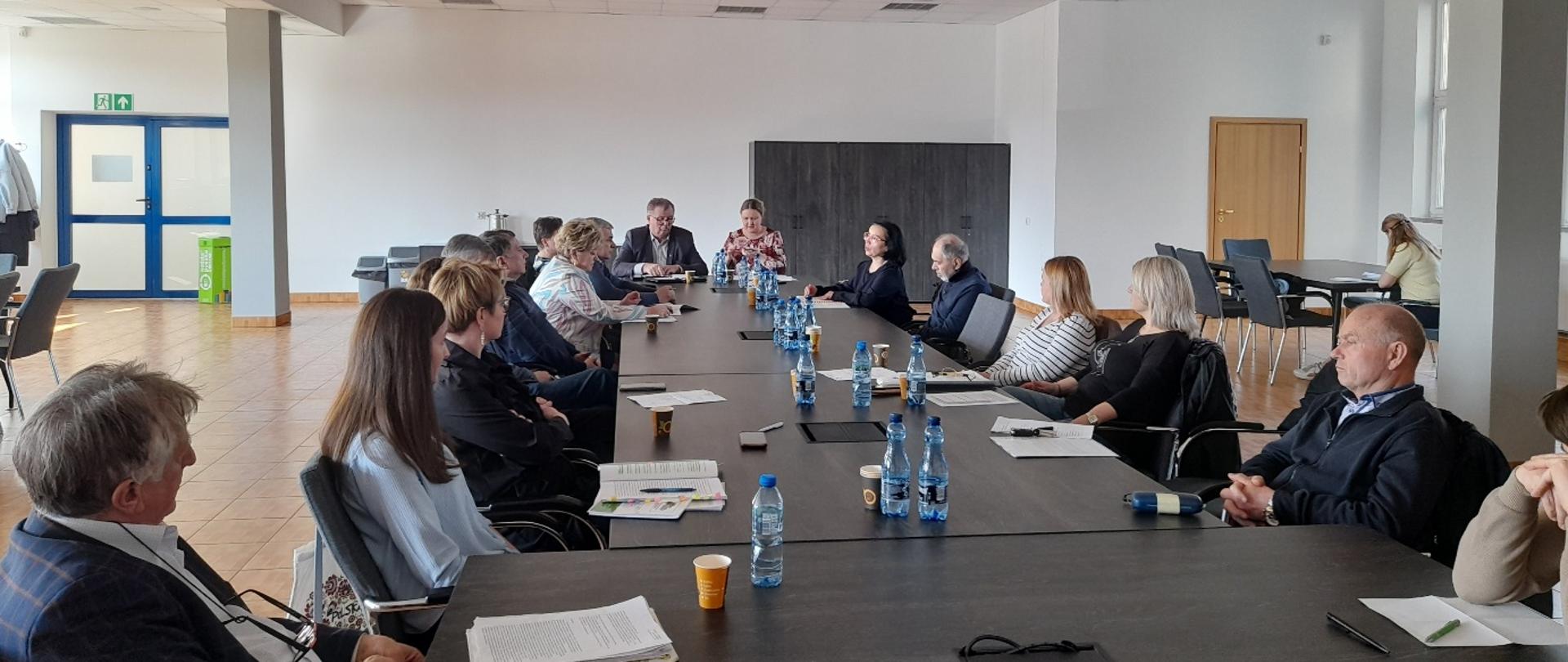 Grupa kilkunastu osób siedzi przy długim stole konferencyjnym w jasnej sali i uczestniczy w spotkaniu; na stole stoją butelki wody i kubki oraz leżą notatki.