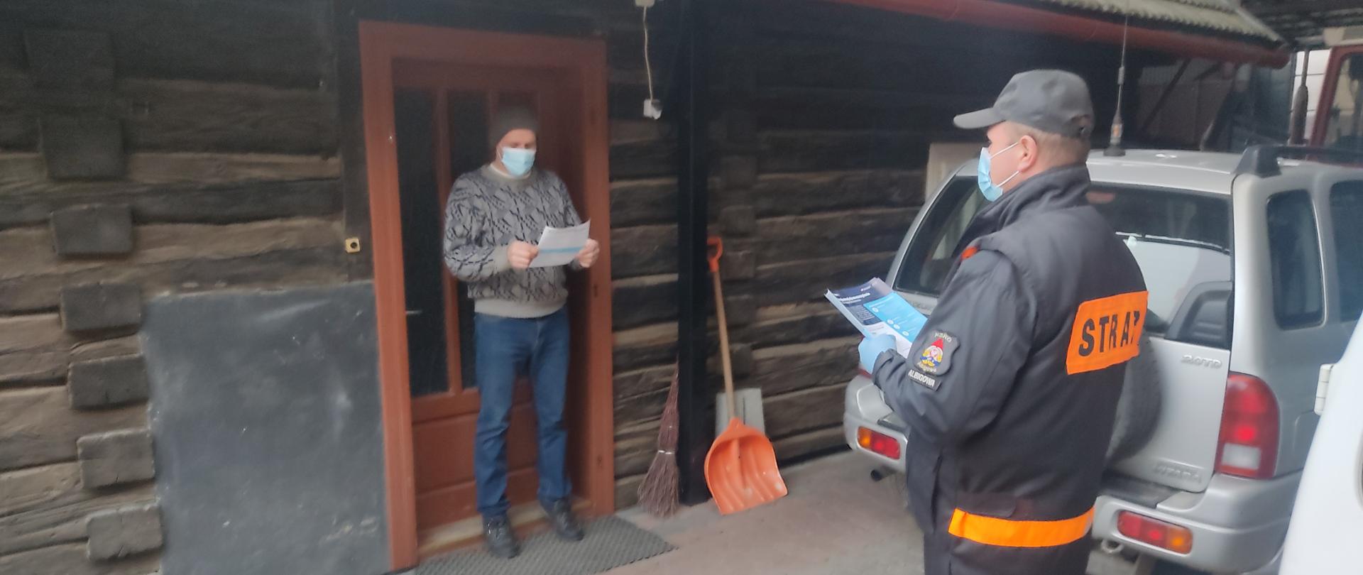 Zdjęcie przedstawia strażaka Ochotniczej Straży Pożarnej w Albigowej trzymającego w ręce ulotki. W tle drewniany dom, w drzwiach, którego stoi mieszkający w nim mężczyzna, który otrzymał ulotkę.