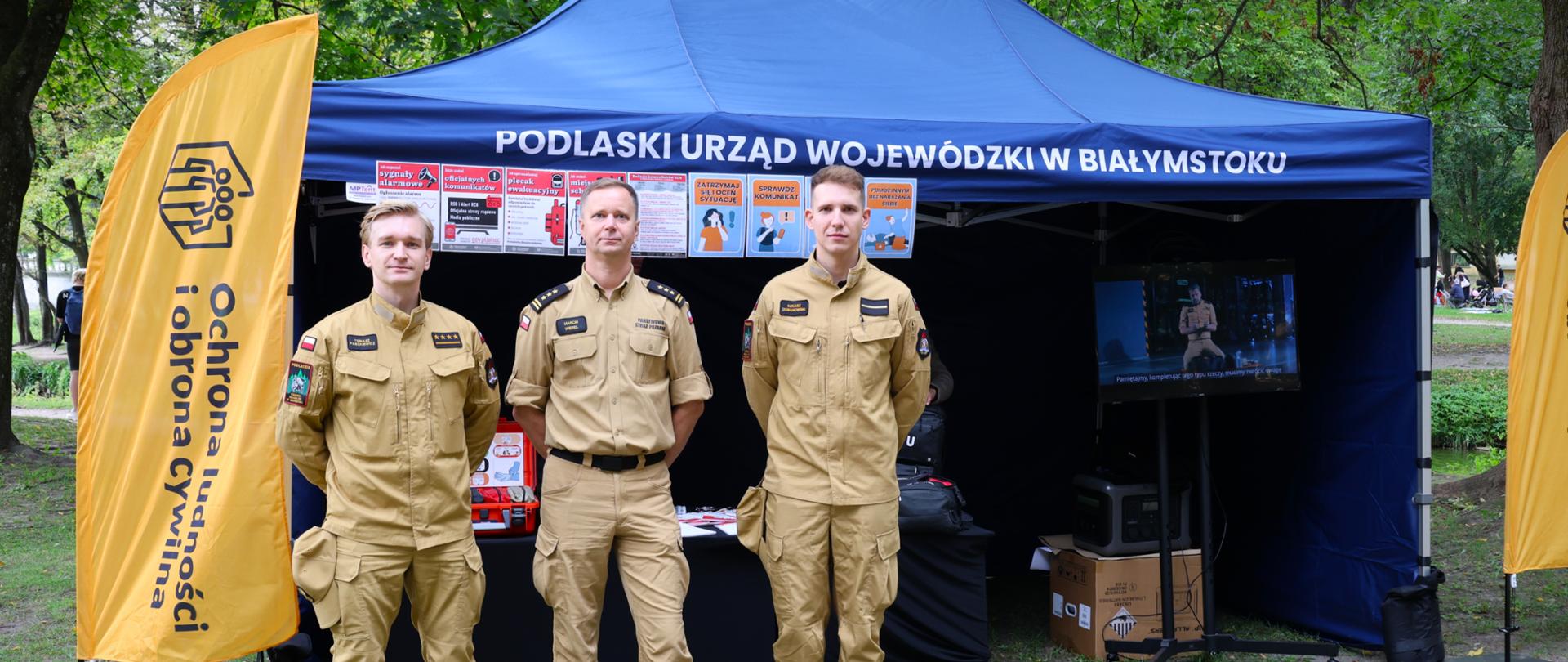  Białostoccy strażacy na czele z zastępcą podlaskiego komendanta wojewódzkiego PSP - st. bryg. Marcinem Wierelem, informowali jak rozpoznawać sygnały alarmowe i prawidłowo na nie reagować - trzech strażaków na tle namiotu OLiOC