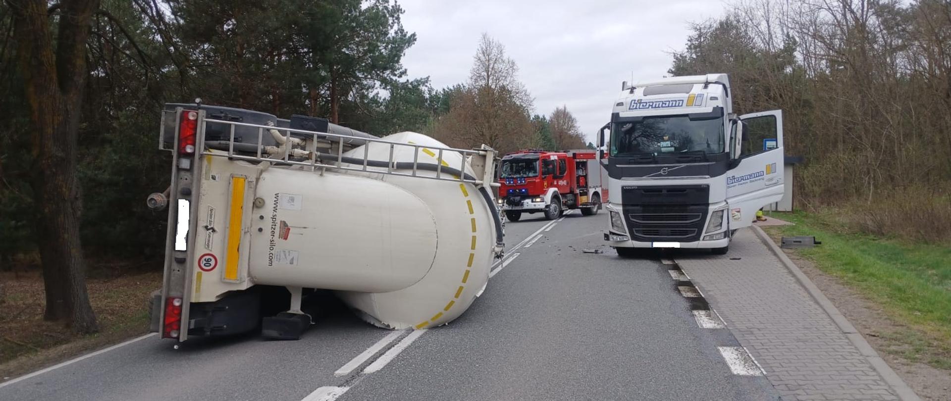 Na drodze leży przewrócona cysterna blokująca oba pasy ruchu. Obok stoi ciężarówka z otwartymi drzwiami, a w tle widać wóz strażacki. Droga przebiega przez zalesiony teren, a na jezdni widoczne są rozrzucone fragmenty po wypadku.
