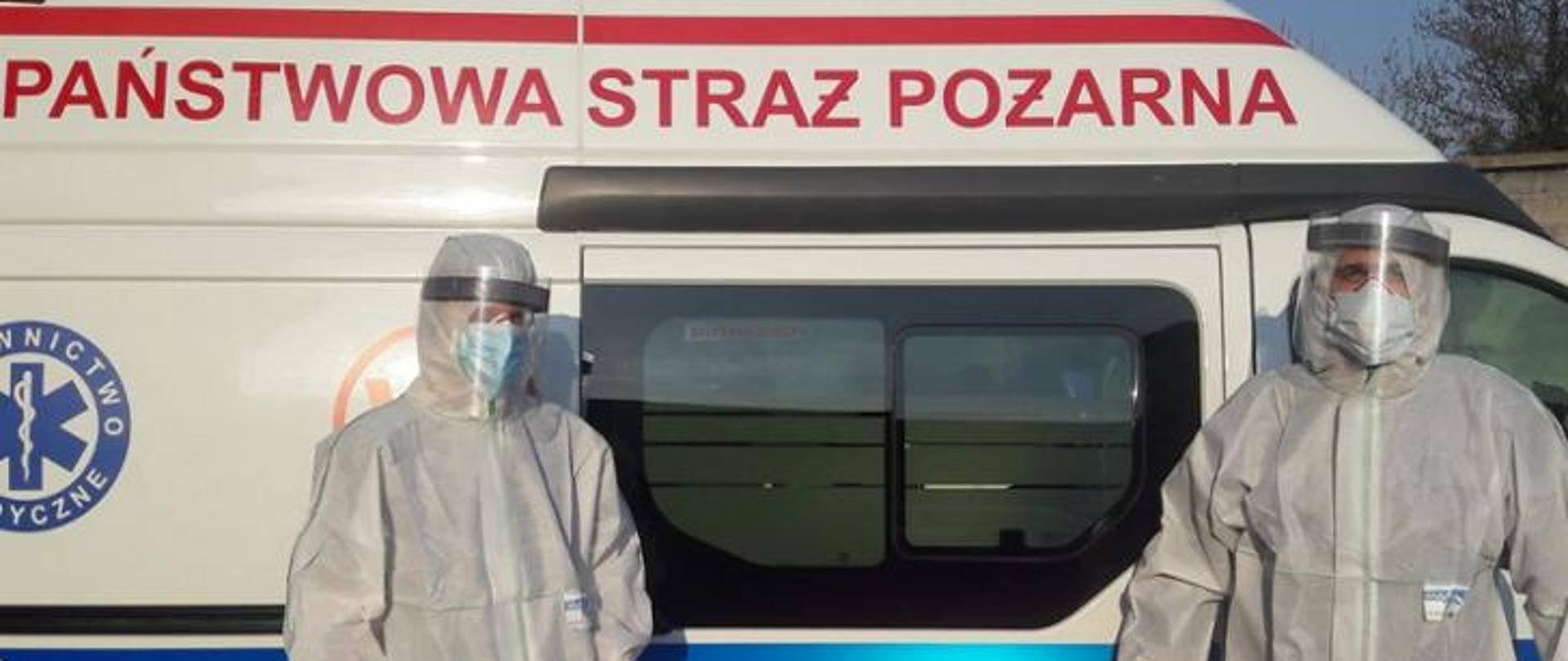 Na tle karetki pogotowia stoi dwóch strażaków zabezpieczonych w ubrania ochronne przeciw COVID-19. Strażacy mają kombinezon ochronny, maseczki i przyłubicę.