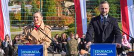 Minister Edukacji i Minister Obrony Narodowej stoją przy dwóch mównicach, na których widnieje napis "Edukacja z wojskiem". W tle widać grupę dzieci i żołnierzy w ramach trwającej lekcji edukacji z wojskiem.
