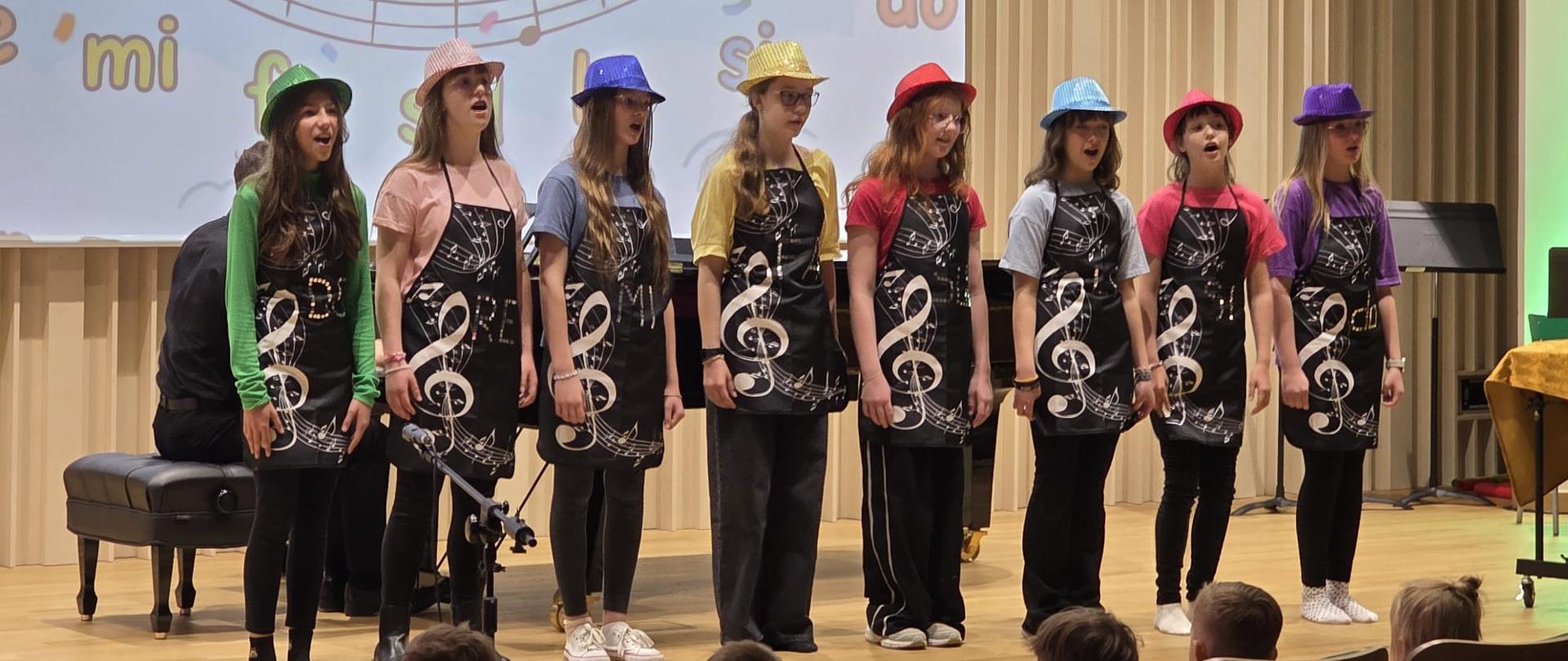 Na scenie sali koncertowej stoi grupa ośmiu dziewczynek podczas wspólnego występu wokalnego. Dzieci ubrane są w kolorowe kapelusze i koszulki w różnych barwach oraz czarne fartuszki z motywem klucza wiolinowego i nut. W tle widoczna jest projekcja z pięciolinią i kolorowymi nutami. Dziewczynki śpiewają przed publicznością siedzącą na widowni. Po lewej stronie częściowo widoczny jest fortepian i akompaniator.