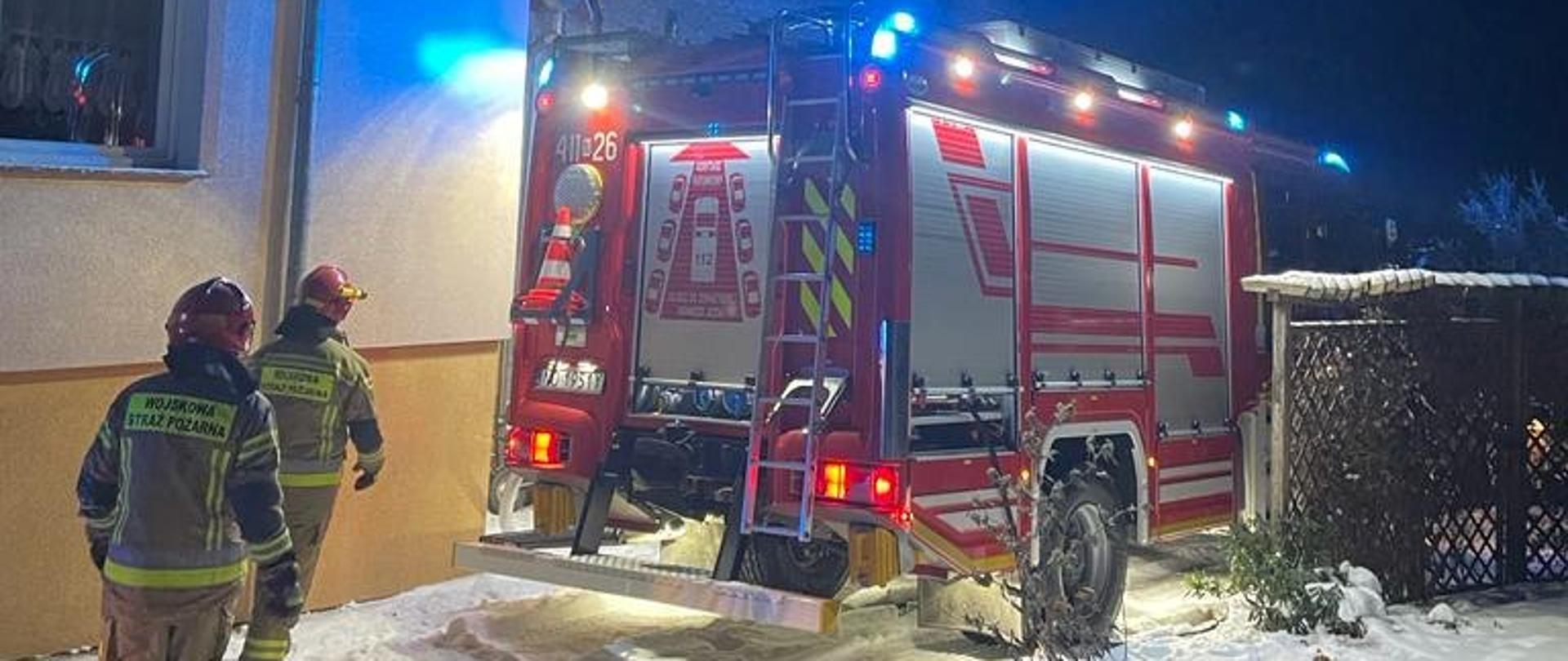 Zdjęcie zrobione nocą. Przy budynku stoi na światłach duży czerwony samochód strażacki. Przy nim strażacy. Biało od śniegu.