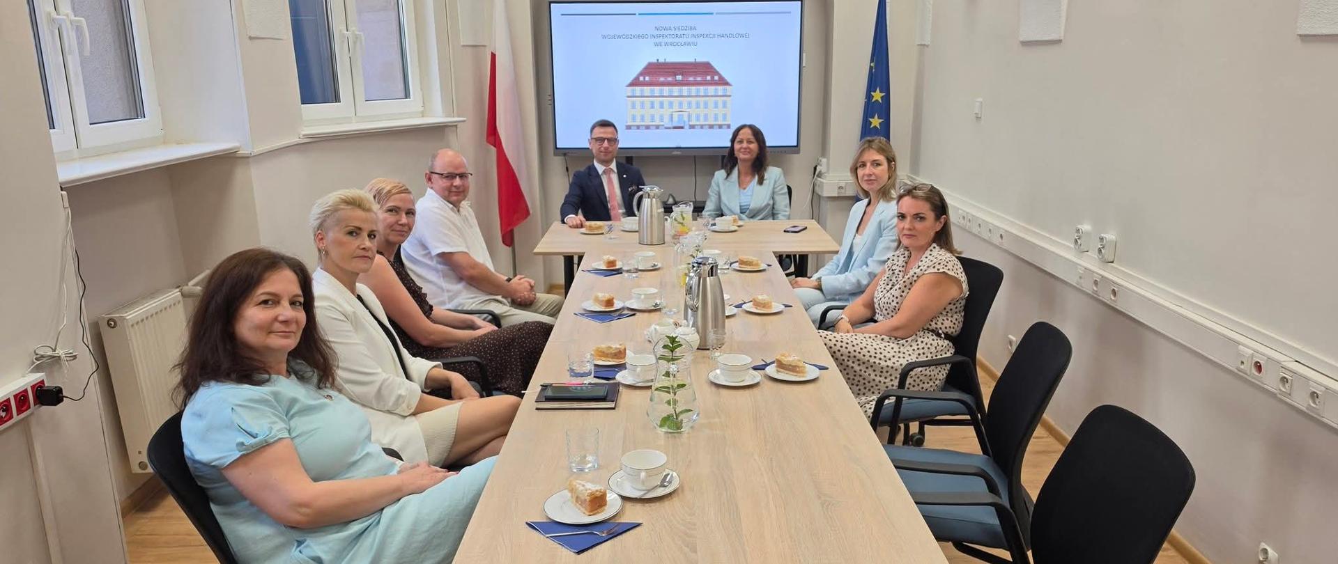Pomieszczenie konferencyjne. Na środku długi stół, po obu stronach siedzą pracownicy Inspekcji Handlowej, na końcu stołu u szczytu siedzi Pani Wicewojewoda i Wojewódzki Inspektor Inspekcji Handlowej. W tle tablica multimedialna z prezentacją, po lewej stronie tablicy stoi flaga Polski, po prawej flaga UE.