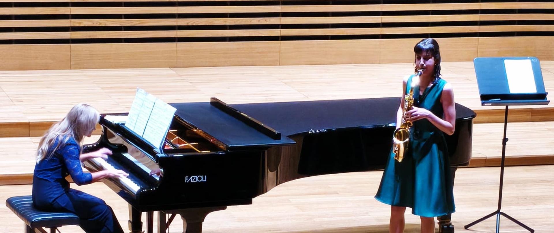 Dziewczyna gra na saksofonie, kobieta akompaniuje na fortepianie.