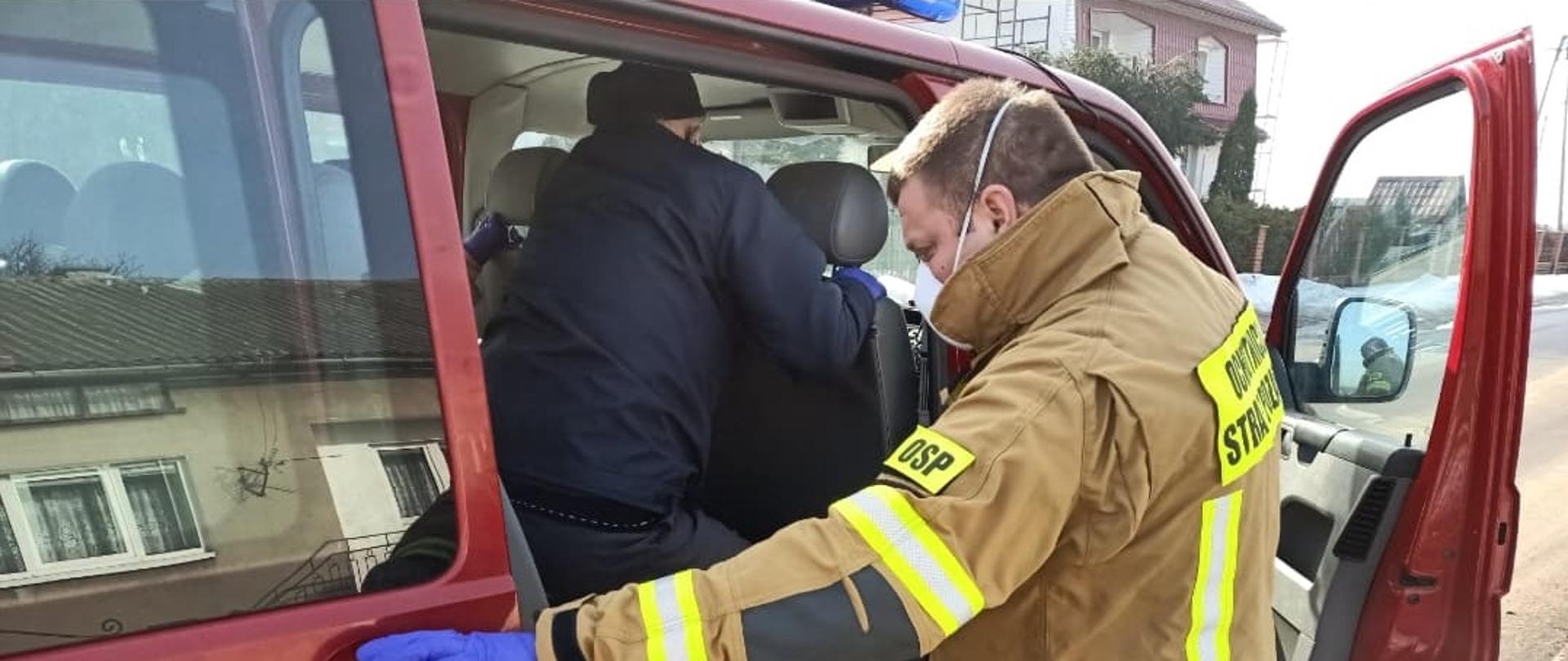 Strażak w żółtym ubraniu specjalnym zamyka drzwi strażackiego samochodu, którym transportuję starszego mężczyznę na szczepienie. 