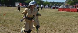 Zawody sportowo-pożarnicze w Jeziorach. Plac ćwiczeń. Trwa jedna z konkurencji zawodów - sztafeta pożarnicza 7x50 m z przeszkodami. Dwie zawodniczki w mundurach koloru piaskowego przekazują sobie pałeczkę sztafetową, którą jest prądownica. Strefa zmian oznakowana jest czerwonymi chorągiewkami. W tle drzewa, przeszkody i kibice.