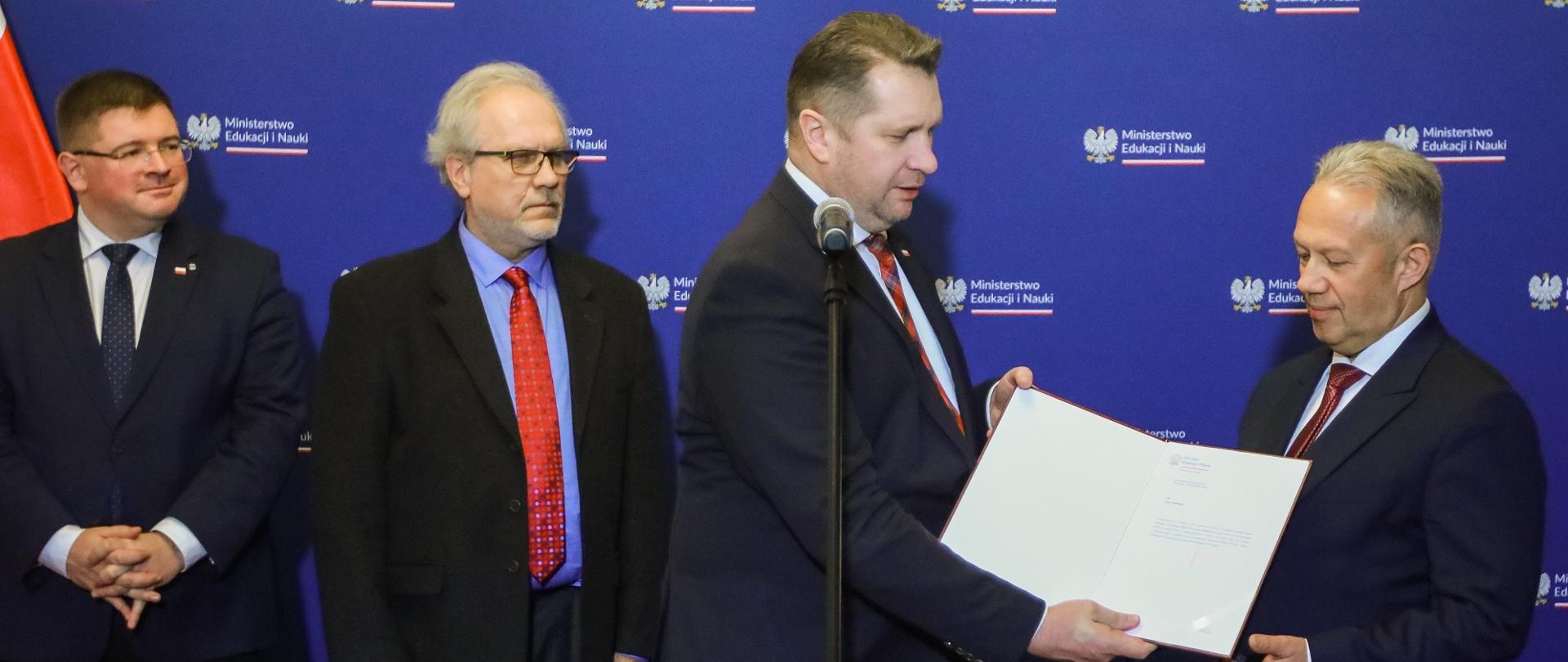 Czterech mężczyzn w garniturach stoi przed niebieską ścianą, wśród nich minister Czarnek który wręcza stojącemu obok otwartą teczkę z dokumentami.