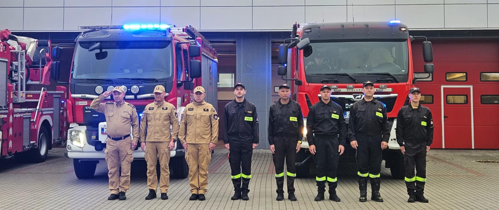Na zdjęciu widać budynek straży pożarnej z dużym napisem „PAŃSTWOWA STRAŻ POŻARNA” nad wjazdami do garaży. Przed budynkiem stoją trzy pojazdy: dwa duże wozy strażackie z włączonymi niebieskimi światłami oraz mniejszy samochód osobowy, policyjny. Na tle budynku i pojazdów ustawiona jest grupa strażaków w mundurach, stojących w szeregu, oddając hołd zmarłemu strażakowi ochotnikowi z Ochotniczej Straży Pożarnej w Michałowie,. Po lewej stronie stoi strażak – dowódca ubrany w mundur służbowy koloru beżowego. Pozostali strażacy siedem osób ubranych w ubrania strażackie koloru czarnego.. Cała scena odbywa się na placu z kostki brukowej, przy jasnym niebie z lekkim zachmurzeniem