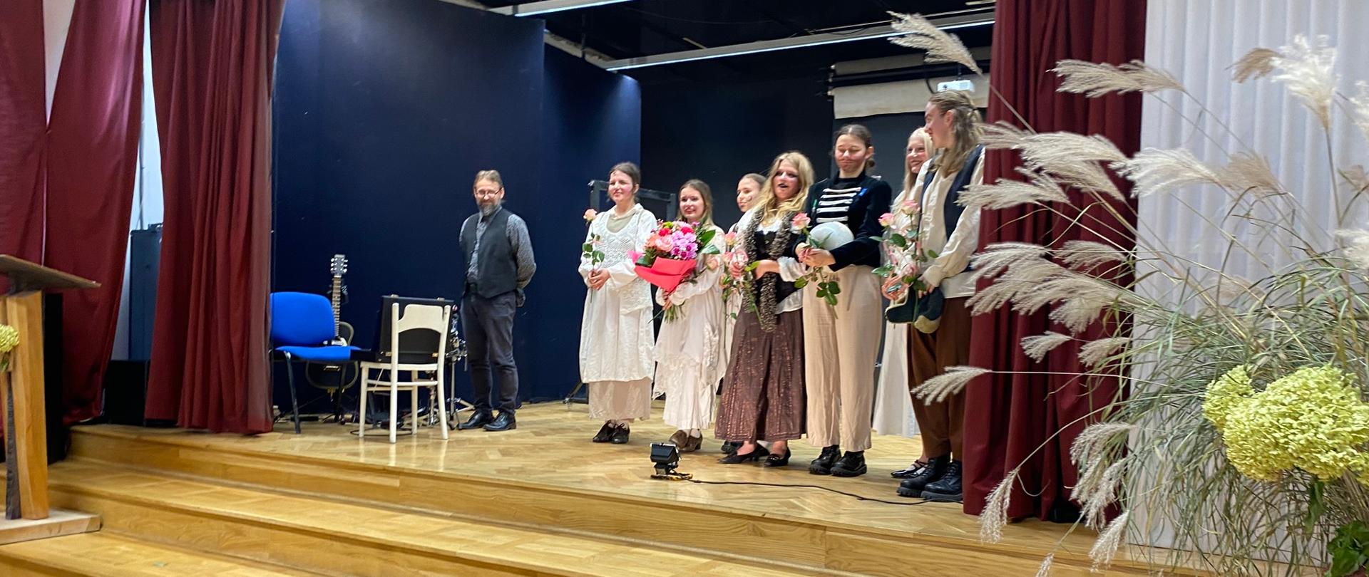 Na drewnianej scenie stoi siedem dziewczyn w strojach scenicznych, wszystkie trzymają kwiaty w dłoniach. Obok nich po prawej stronie stoi mężczyzna, przy nim dwa krzesła, na jednym z nich jest walizka, a z tyłu gitara. Po bokach są bordowe i białe kurtyny. Z prawej stronie jest fragment bukietu z traw i cytrynowych hortensji.