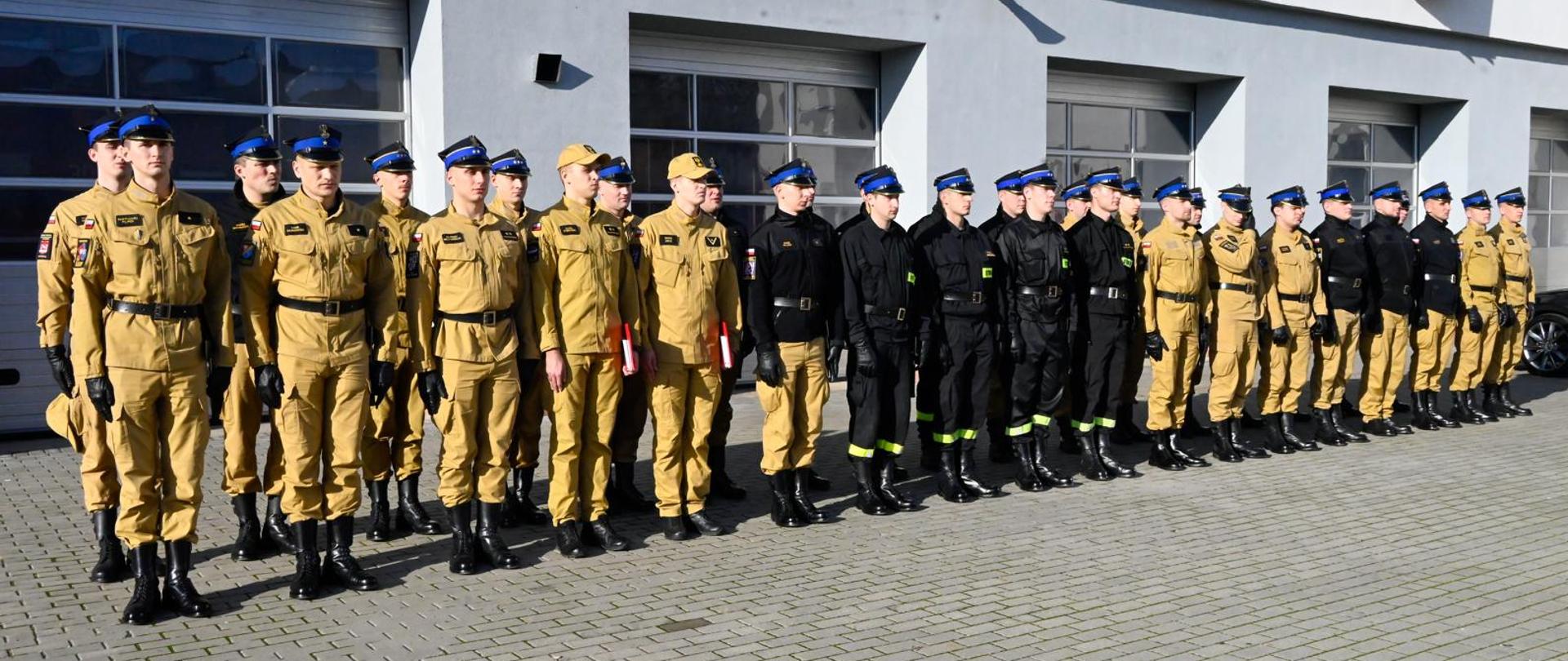 Strażacy PSP w piaskowych mundurach służbowych oraz w czarnych mundurach koszarowych stoją w dwuszeregu. Dodatkowo mają czapki rogatywki. Stoją na parkingu, przed szarym budynkiem garażowym. 