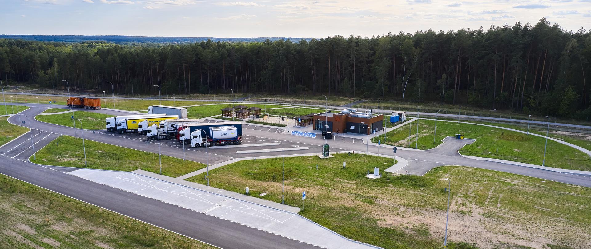 Widok z góry ma miejsce obsługi podróżnych. Widać stojące samochody ciężarowe, budynek z toaletami oraz obok budynku stoliki i miejsca do odpoczynku. Obok miejsca obsługi podróżnych las. 