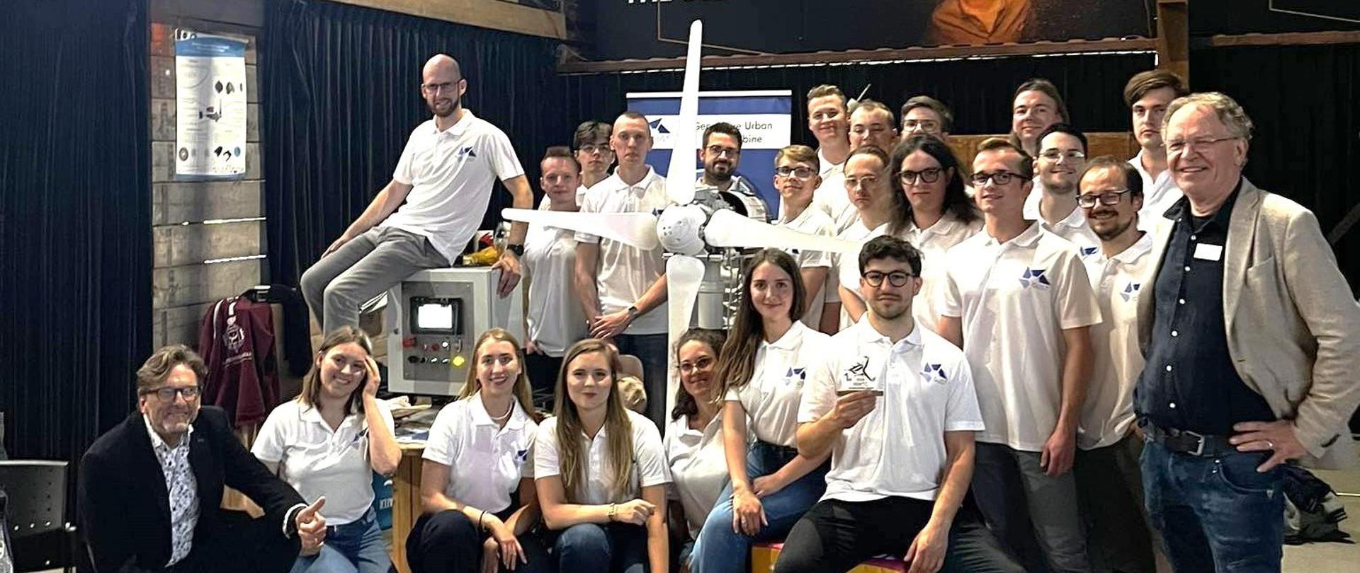 W sali stoi duża grupa młodych ludzi ubranych w białe koszulki, obok stoi mężczyzna w szarej marynarce, kilka osób pośrodku grupy trzyma duże czterołopatowe śmigło.