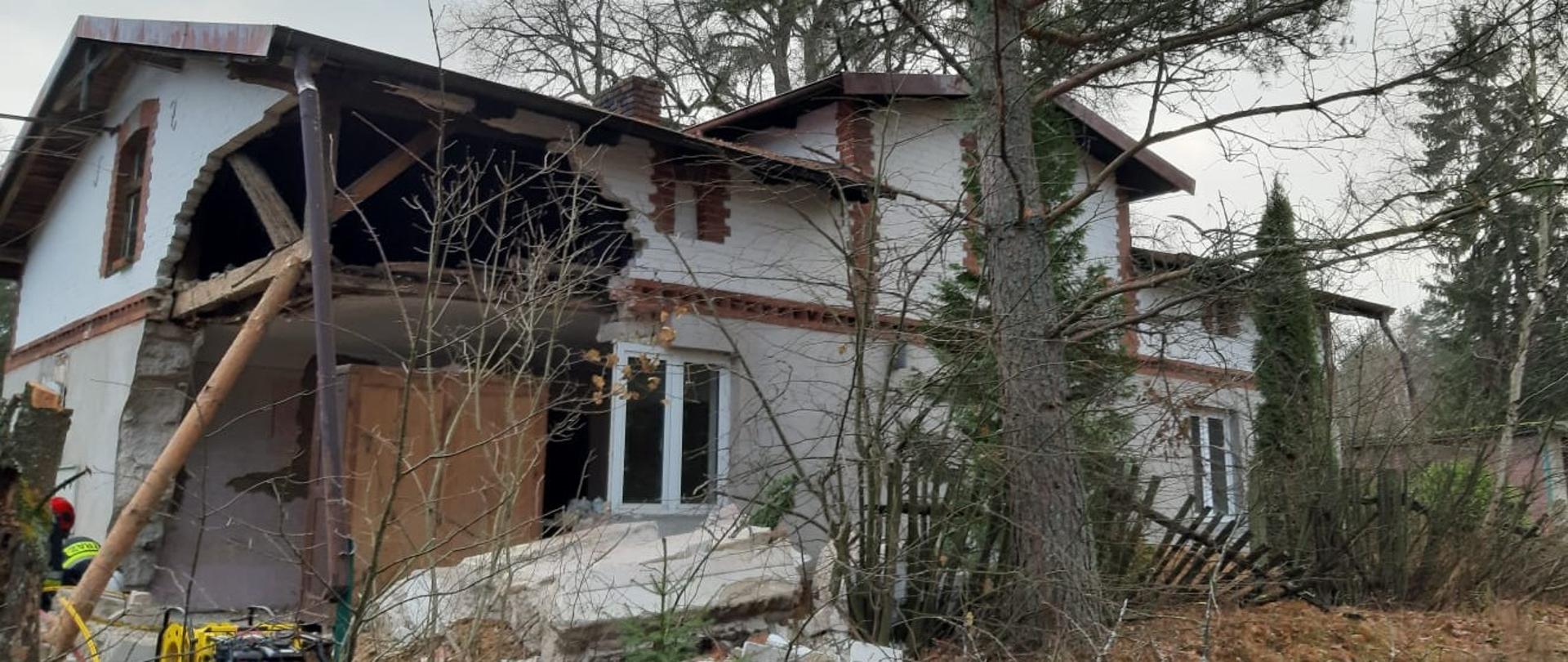 Zdjęcie przedstawia budynek w którym zawaleniu uległa jedna ze ścian, widać gruzowisko, w tle drzewa, widoczny sprzęt ratowniczy PSP