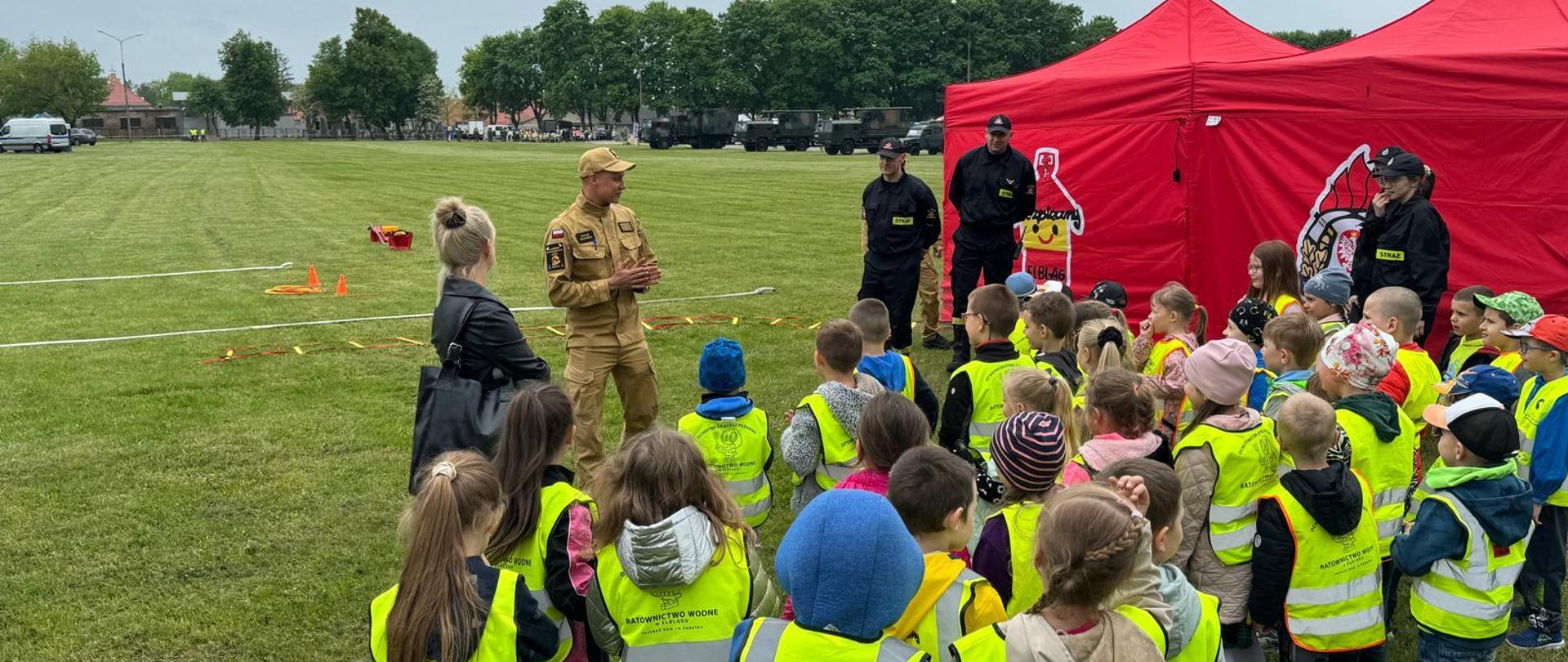 Strażacy w mundurach stoją na tle czerwonego namiotu, otaczają ich z każdej strony dzieci w wieku szkolnym wraz z wychowawczynią. Dzieci mają na sobie żółte, odblaskowe kamizelki.