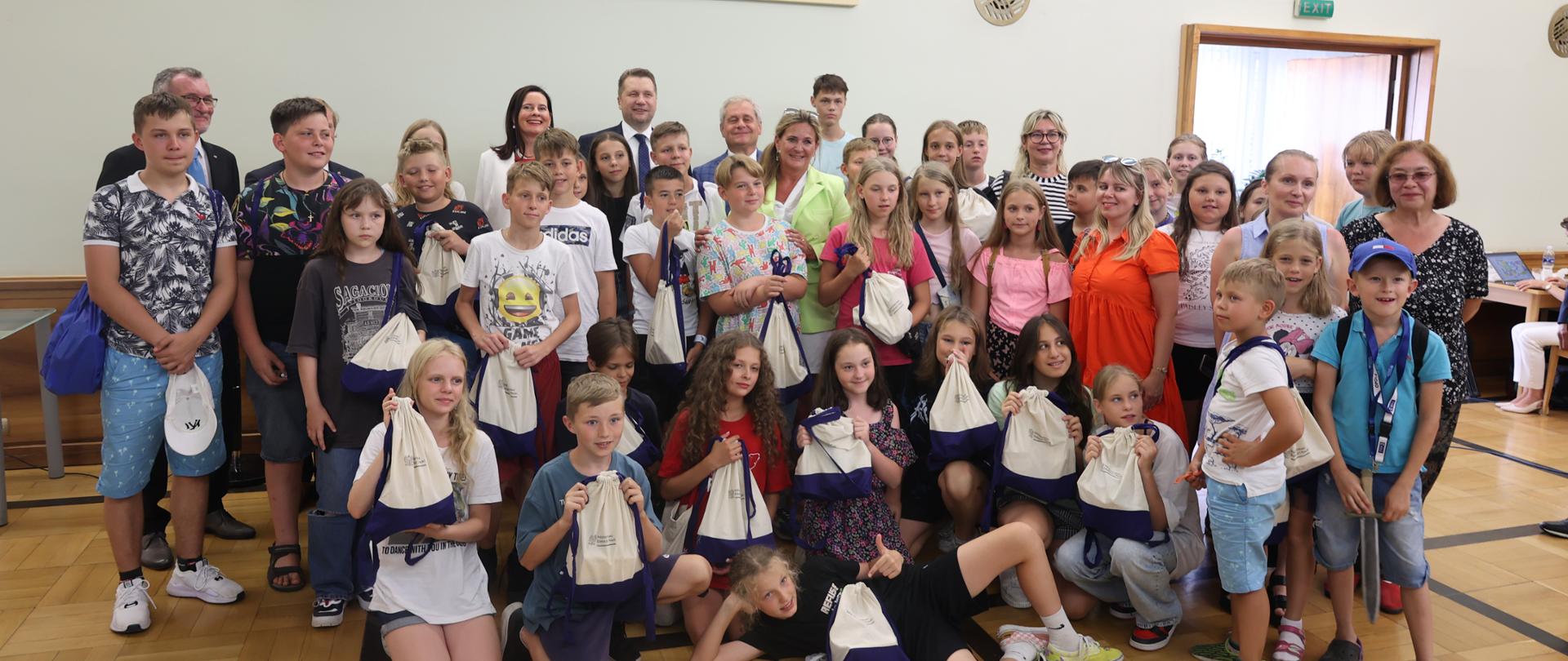 Grupa dzieci wraz z ministrem Czarnkiem i kilkorgiem dorosłych pozuje do zdjęcia, w rekach mają białe woreczki z upominkami. Nad nimi na ścianie duża tablica z napisem: saka im. Prezydenta Lecha Kaczyńskiego.