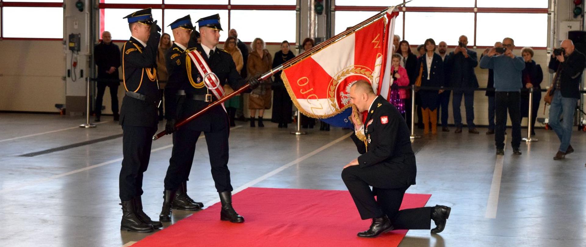 Zdjęcie zrobione w pomieszczeniu, w garażu jednostki ratowniczo-gaśniczej, podczas uroczystego apelu. Poczet sztandarowy trzyma pochylony sztandar. Przed sztandarem klęczy oficer Państwowej Straży Pożarnej w stopniu starszego brygadiera. Strażak całuje materiał sztandaru.